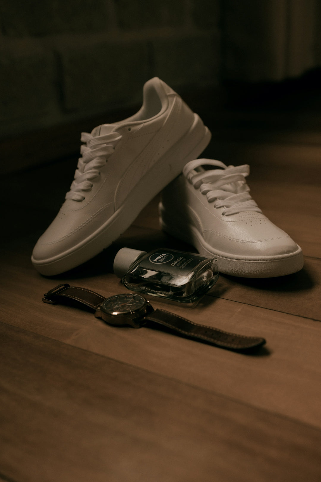 White sneakers, watch, and cologne bottle on wooden floor.